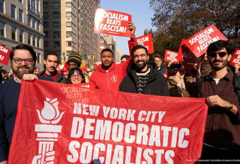 Image of members of NY DSA at a rally holding a banner that says New York City Democratic Socialists. Zohran Mamdani is in the picture as a member of DSA.